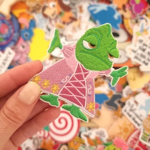 Op de afbeelding: Geborduurde patch met een groene kameleon in een roze jurk met gouden accenten. De patch wordt in een hand gehouden, met een verscheidenheid aan andere kleurrijke patches op de achtergrond. De kameleon heeft een licht chagrijnige uitdrukking.
