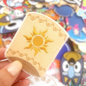 Może przedstawiać: Jasnobrązowa, haftowana naszywka z złotym wzorem słońca. Naszywka ma prostokątny kształt z ozdobnym obramowaniem i jest trzymana na tle innych kolorowych naszywek.