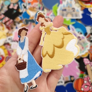 Puede incluir: Parches bordados con Bella de La Bella y la Bestia. Un parche representa a Bella con un vestido azul con una cesta, el otro con un vestido amarillo. Los parches están rodeados de otros parches de personajes.