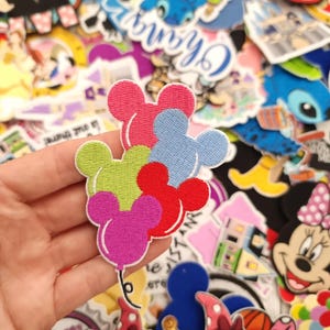 May include: A selection of embroidered patches in various designs. The central patch depicts a cluster of Mickey Mouse-shaped balloons in pink, blue, red, green, and purple. Other patches feature cartoon characters and logos.