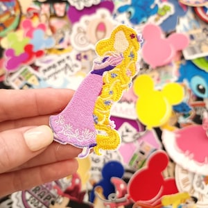 May include: Embroidered patch featuring a cartoon image of a princess with long blonde hair wearing a pink dress. The patch is held in a hand and surrounded by other colorful patches with cartoon characters.