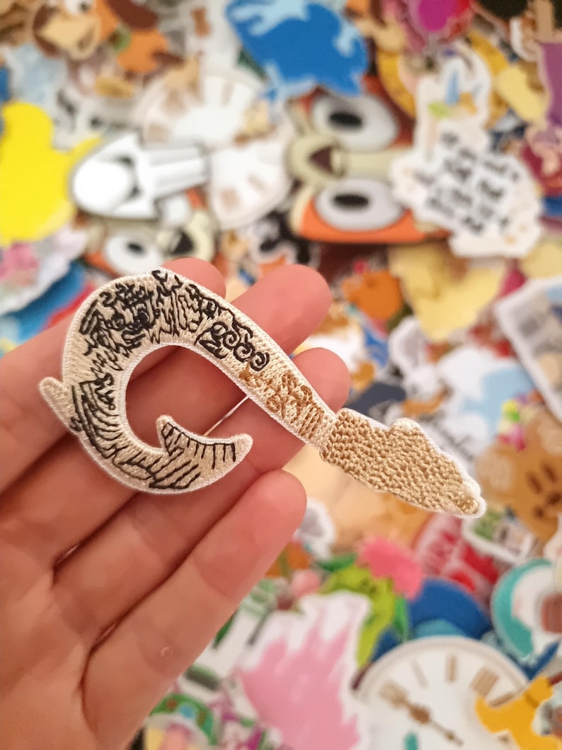 May include: Embroidered patch in the shape of a pirate hook, featuring intricate beige and black detailing. The hook is held against a backdrop of various colorful patches, showcasing a craft or hobby theme.