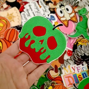 Puede incluir: Un parche verde con forma de manzana con un diseño de calavera en rojo. El parche tiene un tallo negro y detalles rojos goteando. El fondo presenta varios parches y pegatinas coloridos, incluyendo calabazas y personajes de dibujos animados.