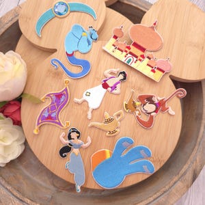 May include: A collection of embroidered patches featuring characters and icons from a classic animated film, including a blue genie, a palace, a magic carpet, a golden lamp, a monkey, and a princess, all arranged on a circular wooden surface.