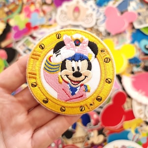May include: Embroidered patch of Minnie Mouse in pink and blue attire, set in a gold circular frame. The patch has a detailed design with a white background, surrounded by other colourful patches.