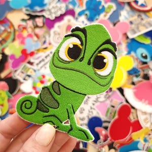 May include: Embroidered patch featuring Pascal, the chameleon from the animated film "Tangled." The patch is primarily green with black and yellow accents, showcasing Pascal's large eyes and expressive features. The patch is surrounded by other colorful patches.