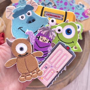 Op de afbeelding: Een verzameling geborduurde stoffen patches met personages en items uit Monsters en Co., waaronder een blauw harig monster, een groen eenogig monster, een bruin monster, een paars wezen in een kostuum en een roze deur met bloemendetails.