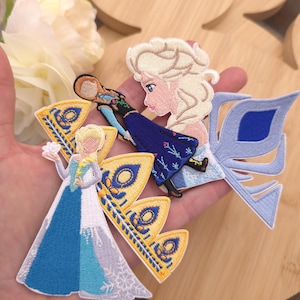 May include: Three embroidered fabric patches featuring animated characters with blonde hair, blue and white gowns, and gold crown accents. The designs include detailed stitching in shades of blue, yellow, and white, showcasing intricate patterns.
