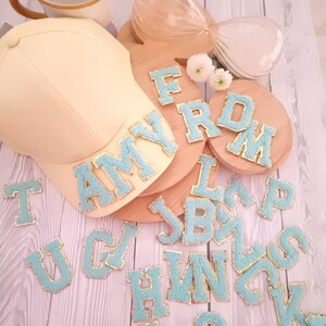 May include: A light yellow baseball cap with the word "AMY" in blue and gold letters on the front.  A wooden board with the word "FROM" in blue and gold letters.  A variety of blue and gold letters are scattered around the cap and board.