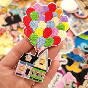 Op de afbeelding: Geborduurde patch met een huis bevestigd aan een tros ballonnen. Het huis heeft een roze, groene en gele gevel met een zwart dak. De ballonnen zijn rood, groen, blauw, geel, paars en roze.