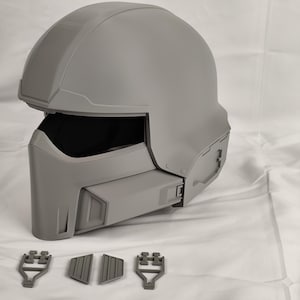 May include: A gray helmet with a visor and a textured surface. The helmet has a futuristic design with multiple details and is made of a hard plastic material. The helmet is accompanied by four small gray plastic pieces.