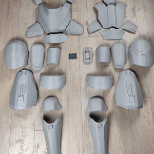 Könnte beinhalten: Zerlegtes graues Kunststoff-Rüstungsset mit Brustplatten, Schulterpolstern, Armschützern, Beinschutz und einem Helm. Die Teile sind auf einer hellen Holzoberfläche angeordnet und zeigen die Komponenten eines Kostüms oder einer Requisite.