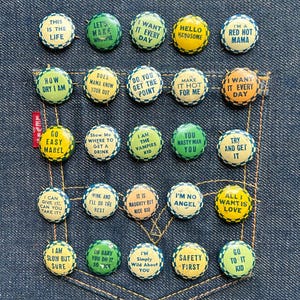 Puede incluir: Una colección de coloridas chapas redondas con varias frases y refranes, dispuestas dentro de un bolsillo de mezclilla. Los botones presentan textos como "This is the Life" e "I'm No Angel."