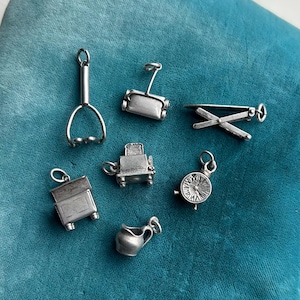 May include: A collection of small, silver-toned charms, including a whisk, a rolling pin, a clothesline, a toaster, a small cabinet, a compass, and a pitcher. The charms are arranged on a teal-colored surface.