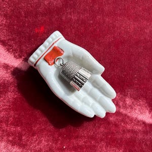 Może przedstawiać: Biała porcelanowa miseczka w kształcie dłoni z czerwoną pieczęcią woskową i srebrną naparstkiem. Naparstek ma wzorzystą fakturę i wisi na małym pierścieniu. Miseczka spoczywa na czerwonej aksamitnej powierzchni.