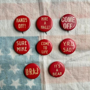 May include: A collection of seven red vintage pinback buttons with white text. The buttons feature phrases like "Hands Off!", "Come Off", and "It's a Bear". The buttons are arranged on a fabric with a blue and white star pattern.