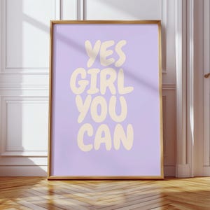 Könnte beinhalten: Ein gerahmtes Poster mit den Worten "YES GIRL YOU CAN" in einer verspielten, cremefarbenen Schrift vor einem hellvioletten Hintergrund. Der Rahmen hat eine warme Goldfarbe, und das Poster lehnt an einer Wand mit Holzboden.