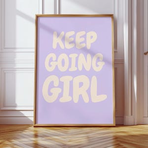 Puede incluir: Una impresión enmarcada con las palabras "KEEP GOING GIRL" en una fuente redondeada de color crema sobre un fondo lila claro. El marco es de color oro cálido. La impresión se muestra sobre un suelo de madera.