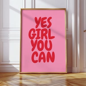 Könnte beinhalten: Ein gerahmter rosa Kunstdruck mit den Worten "YES GIRL YOU CAN" in roter, fetter, handschriftlicher Schrift. Der Druck befindet sich in einem goldenen Rahmen und wird vor einer weißen Wand und einem Holzboden angezeigt.
