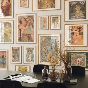 Puede incluir: Una pared de galería con numerosas impresiones artísticas enmarcadas al estilo Art Nouveau con marcos de madera clara. Debajo, una mesa de comedor negra sostiene dos jarrones de vidrio con flores secas, una botella de vidrio pequeña y un libro abierto con páginas blancas.