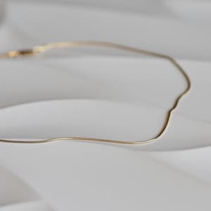 May include: A delicate gold-coloured necklace with a snake chain design. The necklace is displayed on a white surface, with soft shadows adding depth. The chain appears to be approximately 40 cm long.