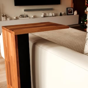 Puede incluir: Una mesa auxiliar de madera con una base de metal negro y una tapa rectangular. La mesa está junto a un sofá blanco con almohadas blancas.