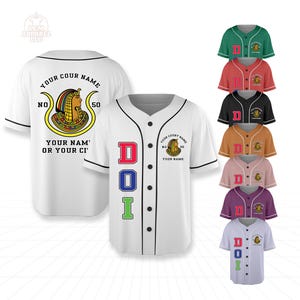 Könnte beinhalten: Weißes Baseball-Trikot mit schwarzem Besatz, mit einem farbenfrohen ägyptischen Emblem und dem Text "YOUR COURT NAME". Das Trikot zeigt die Buchstaben "DOI" in Rot, Blau und Grün. Zusätzliche Trikots in verschiedenen Farben sind ebenfalls abgebildet.
