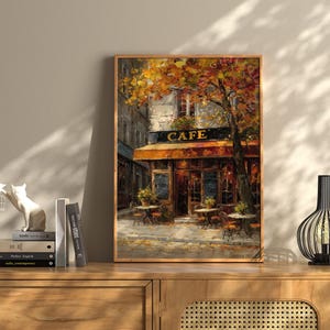 May include: A framed painting of a cafe with the word "CAFE" above the entrance. The cafe has a brown awning and tables outside. The painting is in a wooden frame and is displayed on a wooden cabinet.