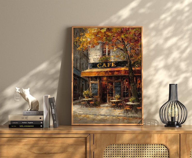 May include: A framed painting of a cafe with the word "CAFE" above the entrance. The cafe has a brown awning and tables outside. The painting is in a wooden frame and is displayed on a wooden cabinet.