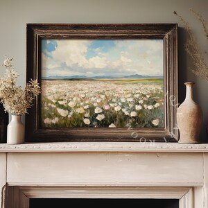 May include: A framed painting of a field of white flowers with a blue sky and white clouds in the background. The painting is hanging on a white mantelpiece above a fireplace.