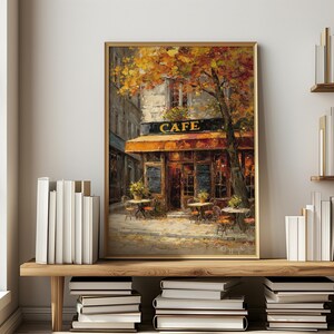 May include: A framed painting of a cafe with an orange awning and the word "CAFE" in gold lettering. The painting depicts outdoor tables and chairs, a tree with yellow leaves, and a cobblestone street. The artwork is displayed on a wooden shelf.