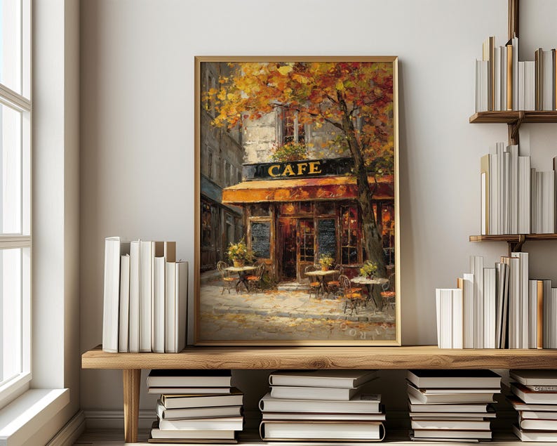 May include: A framed painting of a cafe with an orange awning and the word "CAFE" in gold lettering. The painting depicts outdoor tables and chairs, a tree with yellow leaves, and a cobblestone street. The artwork is displayed on a wooden shelf.