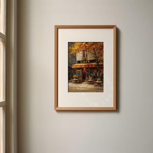 May include: Framed art print of a cafe scene. The artwork depicts a cafe with a brown awning and the word "CAFE" above the entrance. Tables and chairs are set outside. The scene is painted in warm tones, with autumn leaves visible.