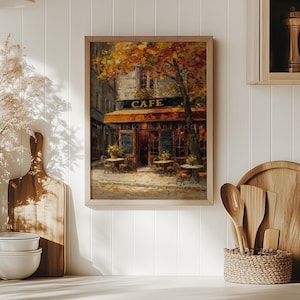 May include: A framed painting of a cafe scene. The cafe has a brown awning and the word "CAFE" above the entrance. Tables and chairs are set up outside. The painting is in a wooden frame and depicts an autumn scene.