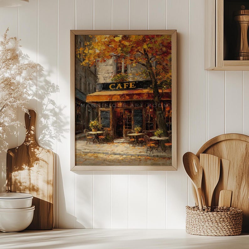 May include: A framed painting of a cafe scene. The cafe has a brown awning and the word "CAFE" above the entrance. Tables and chairs are set up outside. The painting is in a wooden frame and depicts an autumn scene.