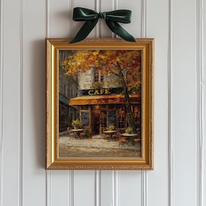 May include: A framed painting of a cafe scene with a gold ornate frame. The painting depicts a cafe with outdoor seating, a sign that reads "CAFE", and autumn foliage. A dark green velvet bow is tied at the top of the frame.
