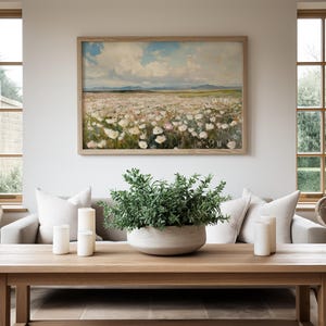 May include: A living room with a white sofa, two wicker chairs, and a wooden coffee table. The coffee table has three white candles and a green plant in a gray bowl. A framed painting of a field of white flowers hangs on the wall.