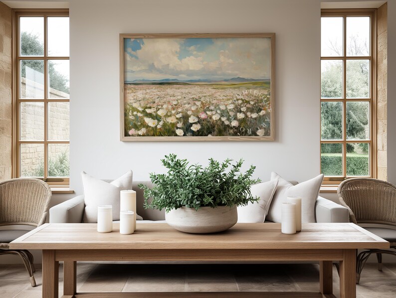 May include: A living room with a white sofa, two wicker chairs, and a wooden coffee table. The coffee table has three white candles and a green plant in a gray bowl. A framed painting of a field of white flowers hangs on the wall.