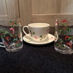 May include: A festive Christmas tea set. Two clear glass mugs with handles, decorated with candy canes, holly, and berries. A white teacup and saucer feature similar festive designs, perfect for the holiday season.