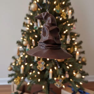 May include: A brown Sorting Hat Christmas tree decoration hangs in front of a decorated Christmas tree. The ornament is a replica of the hat from the Harry Potter series. The tree is adorned with lights, ornaments, and candles, with wrapped gifts at the base.