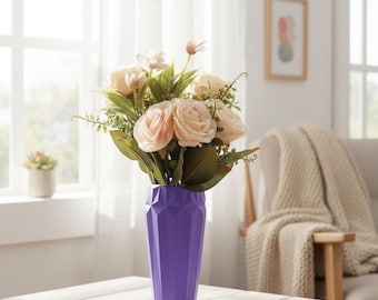 May include: A purple geometric vase holding a bouquet of peach-colored roses and greenery. The vase sits on a white wooden table. The scene is set in a well-lit room with a window and a chair in the background.