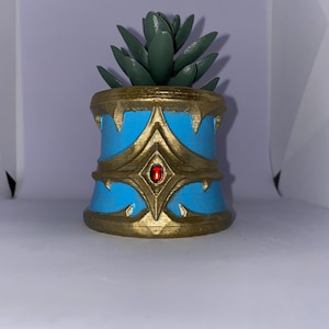 May include: A decorative planter with a succulent plant. The planter is cylindrical, with a light blue base and gold accents. A red gem is set in the center of the gold design. The succulent has green leaves.