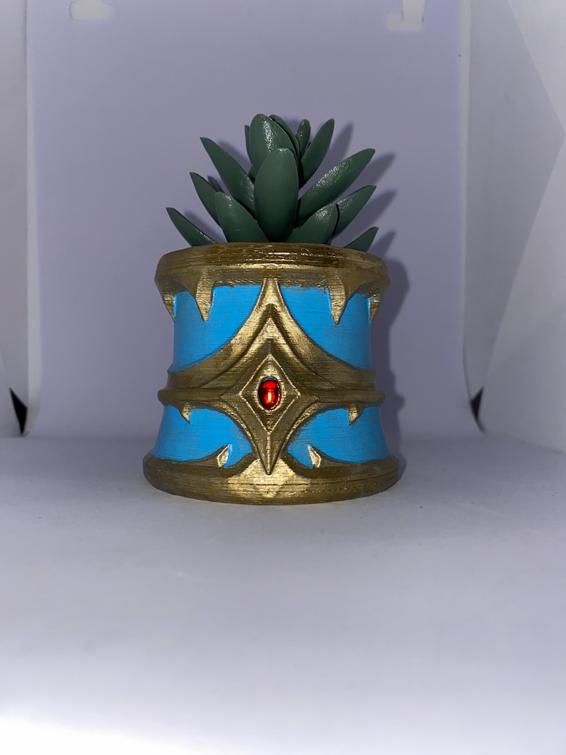 May include: A decorative planter with a succulent plant. The planter is cylindrical, with a light blue base and gold accents. A red gem is set in the center of the gold design. The succulent has green leaves.