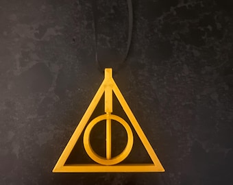 May include: A gold-colored pendant shaped like the Deathly Hallows symbol from the Harry Potter series. The symbol is a triangle with a circle inside and a vertical line through the circle.