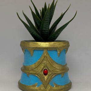 May include: A small, decorative planter with a faux succulent. The planter is cylindrical, painted light blue with gold accents and a red jewel. The succulent has dark green, pointed leaves.