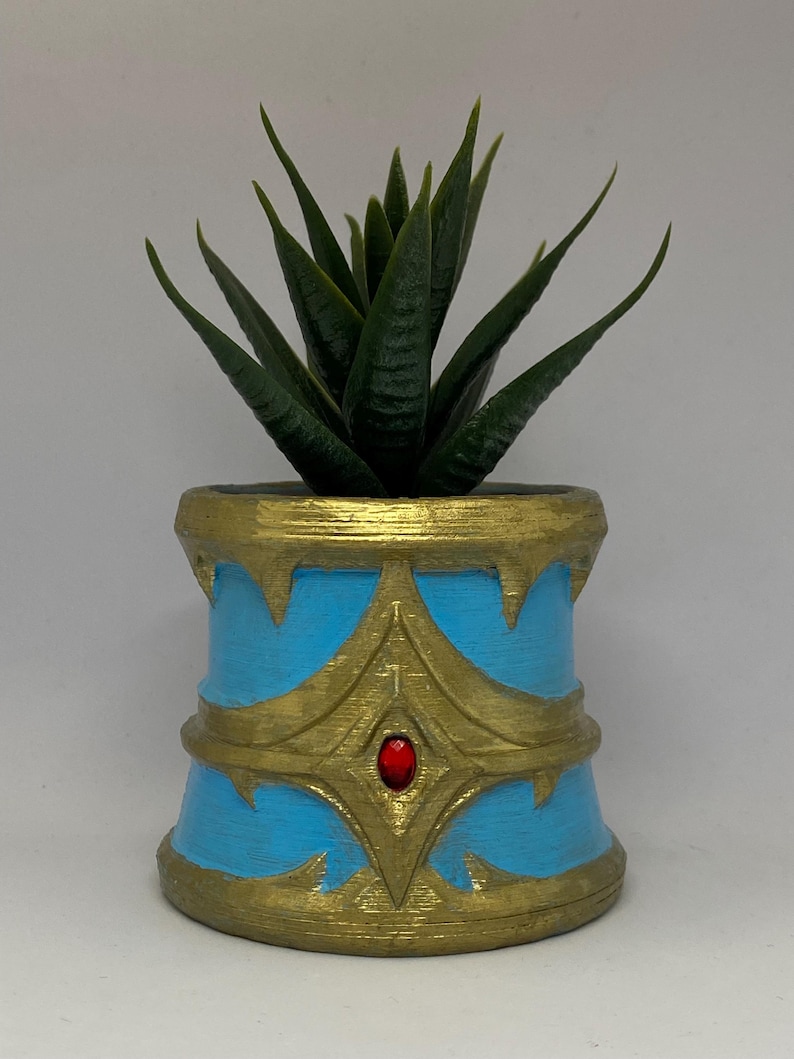 May include: A small, decorative planter with a faux succulent. The planter is cylindrical, painted light blue with gold accents and a red jewel. The succulent has dark green, pointed leaves.