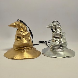 May include: Two hanging ornaments shaped like pointed, folded hats. One ornament has a metallic gold finish, and the other has a metallic silver finish. Each hat features a wide brim, a creased crown, and a black ribbon loop attached to the top.