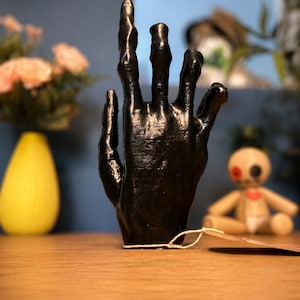 May include: A black, upright, sculpted hand figurine with elongated, textured fingers. The hand is positioned vertically with a thin, light-colored string tied around the base, attached to a small, blank, light brown paper tag extending to the right.