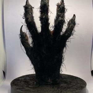 Può includere: Una scultura nera a forma di mano di creatura a cinque dita con lunghi artigli metallici curvi e affilati. La mano è montata in posizione verticale su una base circolare nera, caratterizzata da una superficie ruvida e pelosa.