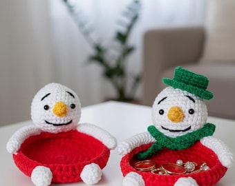 Könnte beinhalten: Zwei handgefertigte Schneemann-Schmuckschalen aus Häkelarbeit. Die Schneemänner haben weiße Köpfe, rote Körper und kleine weiße Füße. Einer der Schneemänner trägt einen grünen Hut und Schal. Die Schalen sind mit Schmuck gefüllt, darunter Ringe und Perlen.